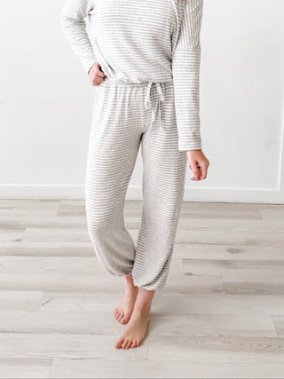 A woman modeling a matching long-sleeve top and jogger pants pajama set in a white and light gray striped pattern, standing in front of a white paneled wall with 'STYLE & SPIRE' text.