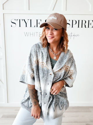 A woman models a gray and white knit cardigan with brown geometric patterns, paired with a brown baseball cap and white pants