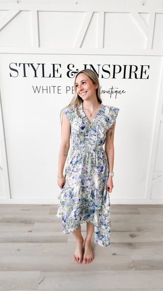 Woman wearing a blue and green floral dress with ruffle sleeves and a high to low detail on the bottom of the dress. 